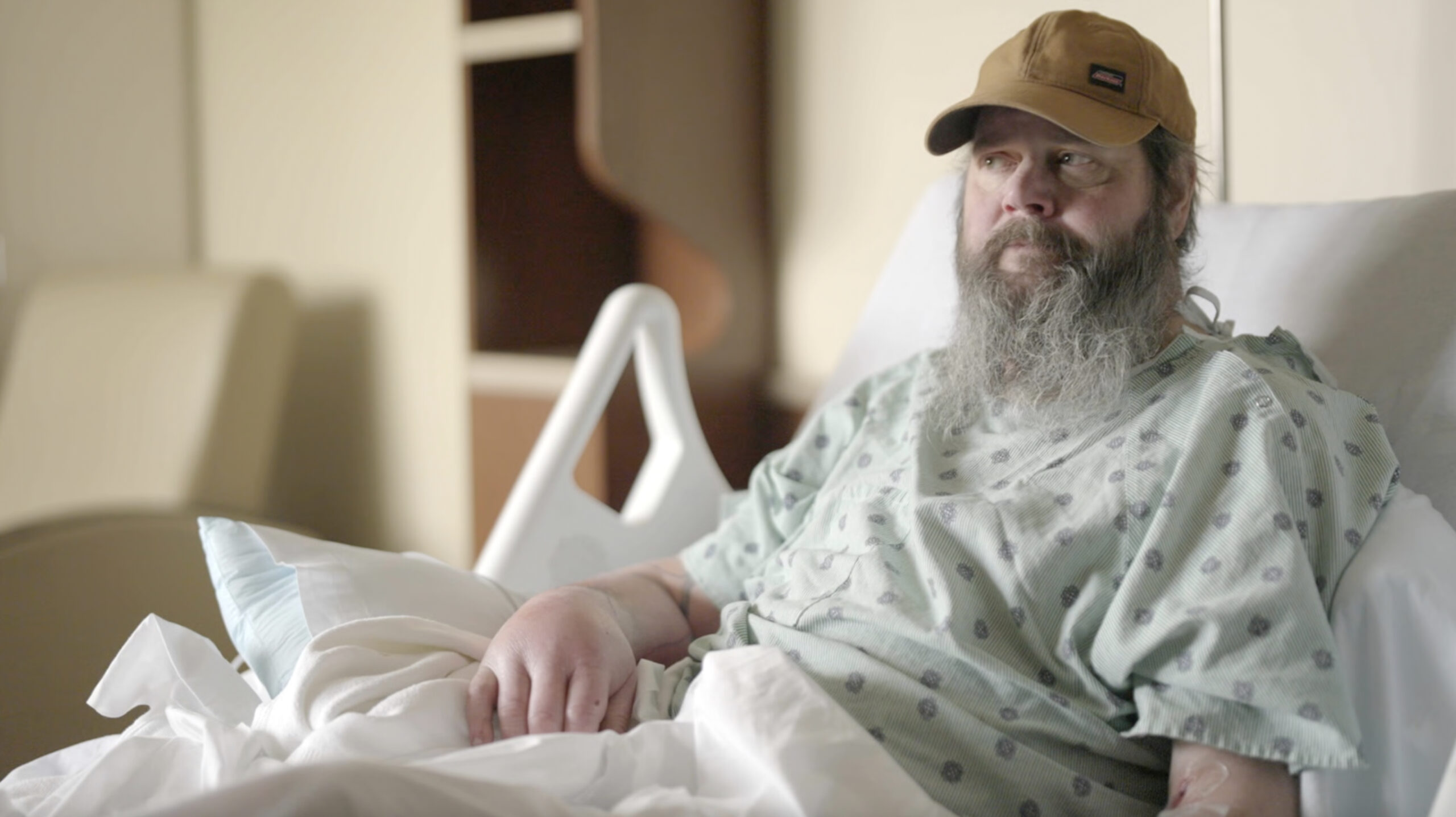 Bearded man in a hospital gown sits up in bed, wearing a tan cap and looking to the right.